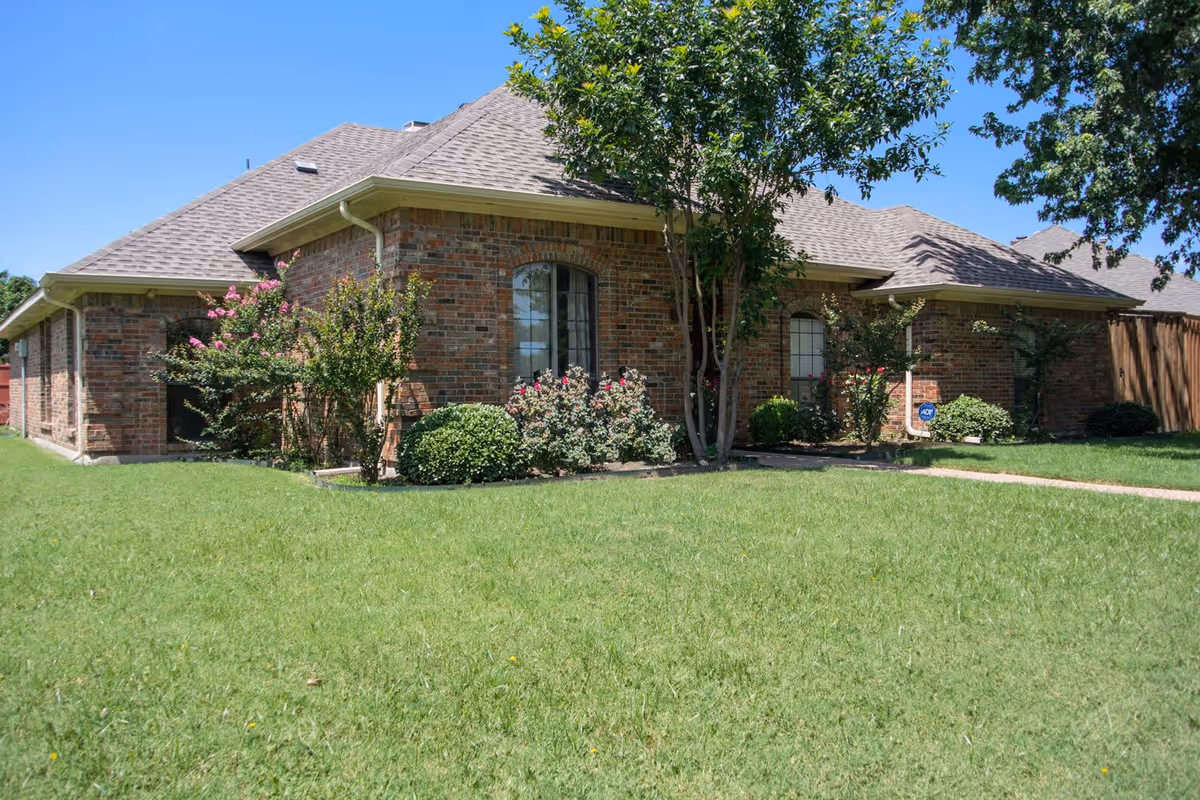 Exterior view of a single-story brick building with a sloped roof, surrounded by green grass and landscaped bushes and trees under a clear blue sky.