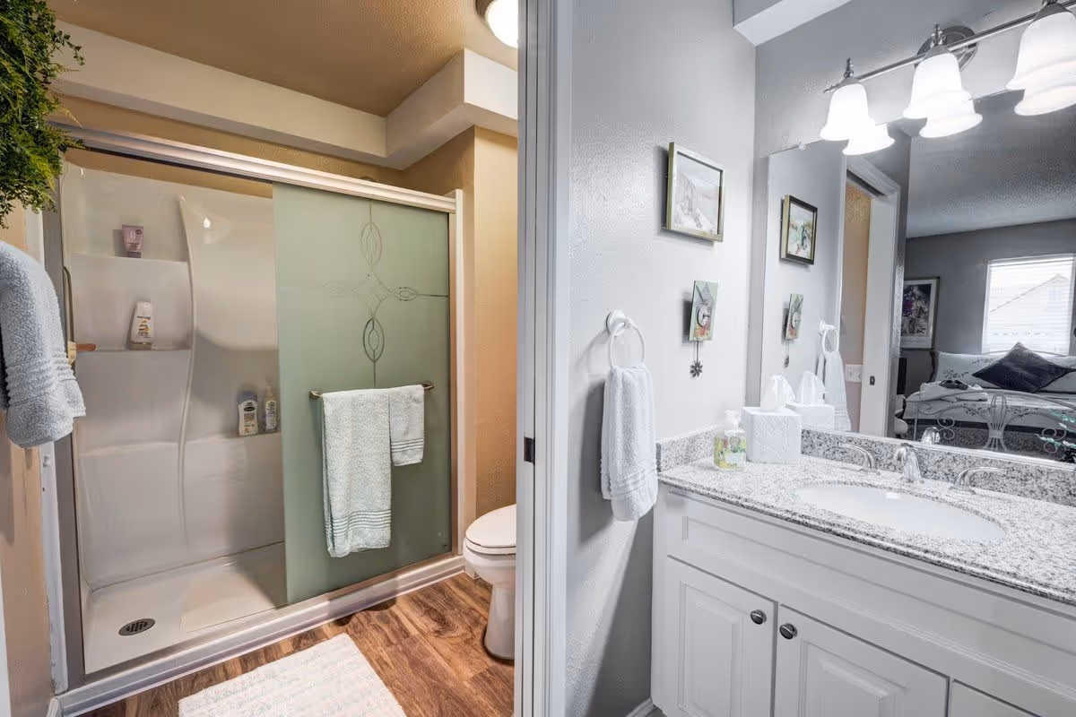 A clean bathroom with a glass sliding shower door featuring a decorative frosted design, a white towel hanging on the door, a toilet next to the shower, and a white vanity with a granite countertop, sink, and a large mirror above. The bathroom has wood-style flooring and soft lighting. A glimpse of a bedroom with a bed and window is visible through the doorway.