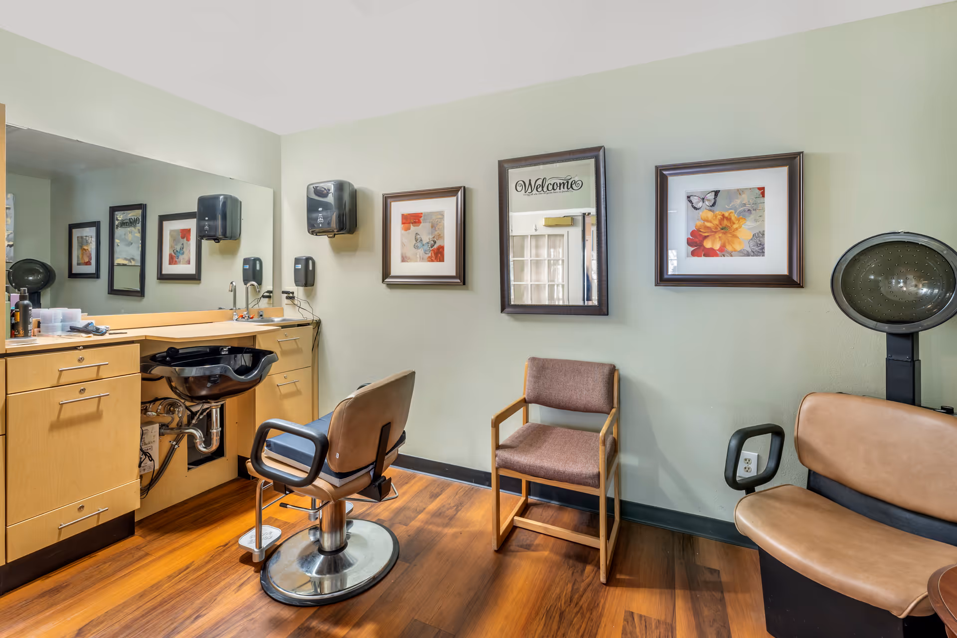 Interior view of a hair salon area in a senior living facility with a salon chair in front of a black hair washing sink, wooden cabinets, a large mirror, two framed floral pictures on the wall, a brown cushioned chair, and a vintage hair dryer chair.