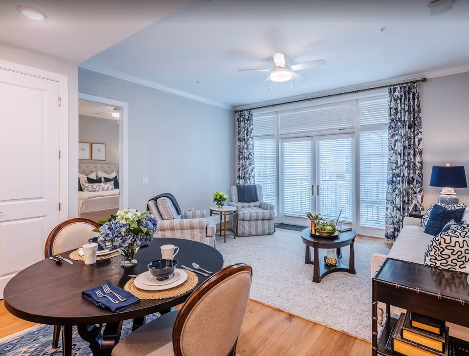A bright and cozy living room with a round dining table set for two in the foreground. The living area features two striped armchairs, a round coffee table with decorative plants, a white sofa with patterned cushions, and a blue lamp on a side table. Large windows with white blinds and floral curtains let in natural light. A doorway leads to a bedroom with a neatly made bed and framed artwork on the wall.