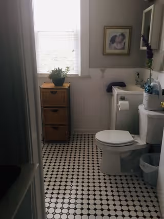 A small bathroom with a white toilet, a white sink with a mirror above it, a wicker drawer unit with a potted plant on top, a window with blinds, a framed picture on the wall, and a tiled floor with a black and white pattern.