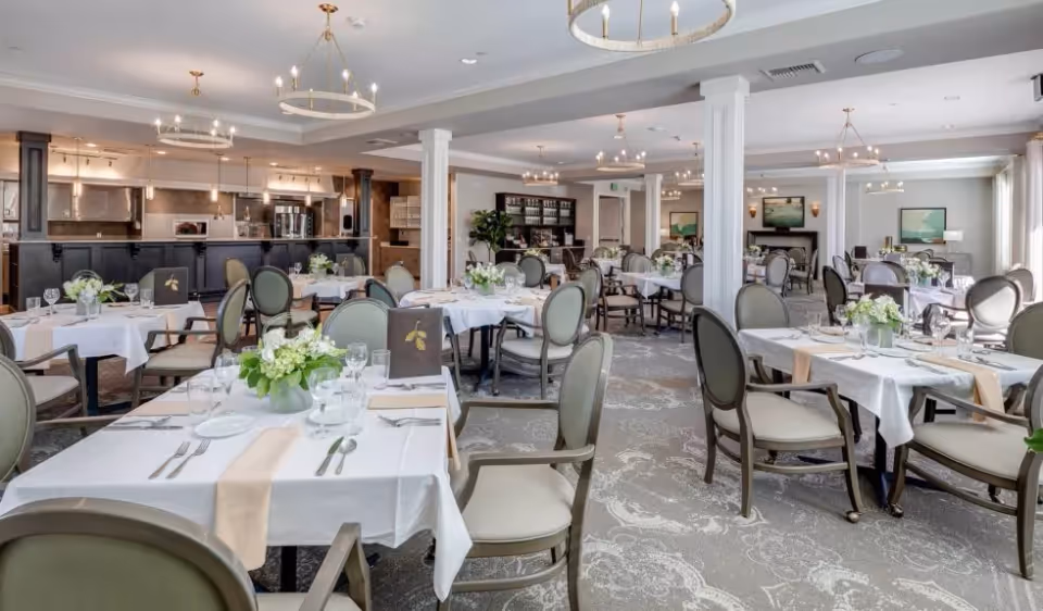 A spacious, elegantly decorated dining room with multiple tables covered in white tablecloths, each set with plates, glasses, silverware, and floral centerpieces. The room features comfortable chairs with armrests, chandeliers hanging from the ceiling, and a kitchen area visible in the background.