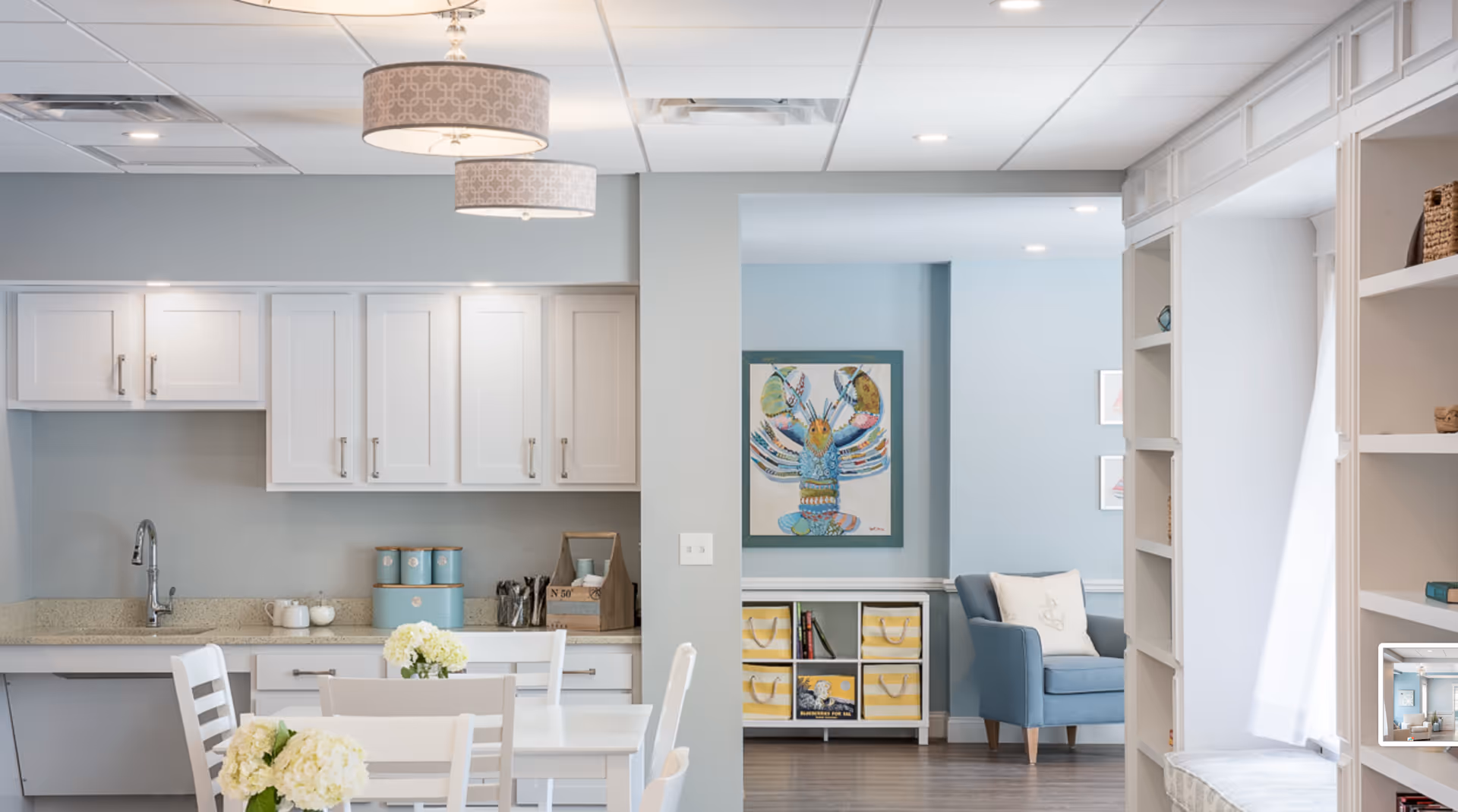 Bright and clean interior space featuring a white kitchen area with cabinets, a sink, and a countertop. In the foreground, there is a white dining table with white chairs and small flower arrangements. Beyond the kitchen, there is a cozy sitting area with a blue armchair, a white storage unit with yellow-striped baskets, and a colorful lobster painting on the light blue wall. Built-in white shelves with decorative items are visible on the right side.