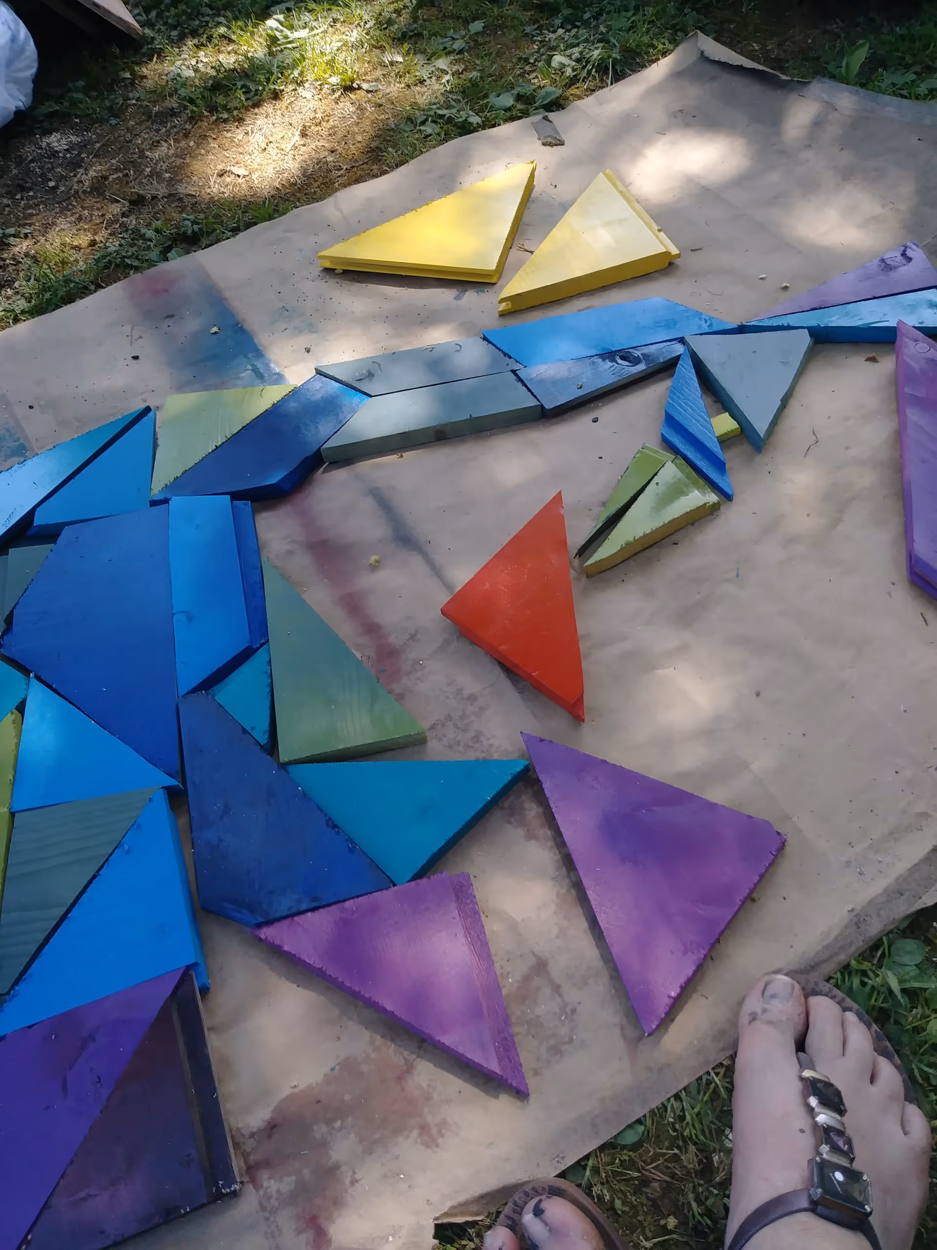 Colorful painted wooden triangular pieces laid out on paper on the grass with a sandal-clad foot visible in the corner.