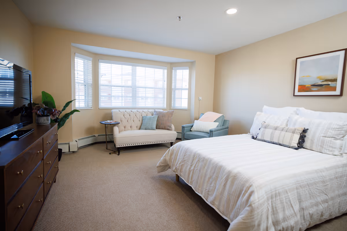 A bright bedroom with a large bed covered in white linens and multiple pillows. There is a light blue armchair with a white pillow and a pink throw blanket next to a small round side table. A tufted beige loveseat with two pillows is positioned in front of a large bay window with white blinds. A wooden dresser with a flat-screen TV and a potted plant is on the left side of the room. A framed abstract painting hangs on the beige wall above the bed.