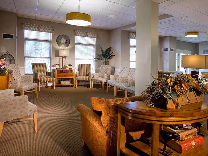 A spacious and well-lit living room area with multiple upholstered chairs arranged in a semi-circle near windows with blinds. There is a wooden side table with a lamp and decorative items, a large clock on the wall, and a wooden console table with a floral arrangement and books. The room has beige walls, carpeted floor, and ceiling lights.