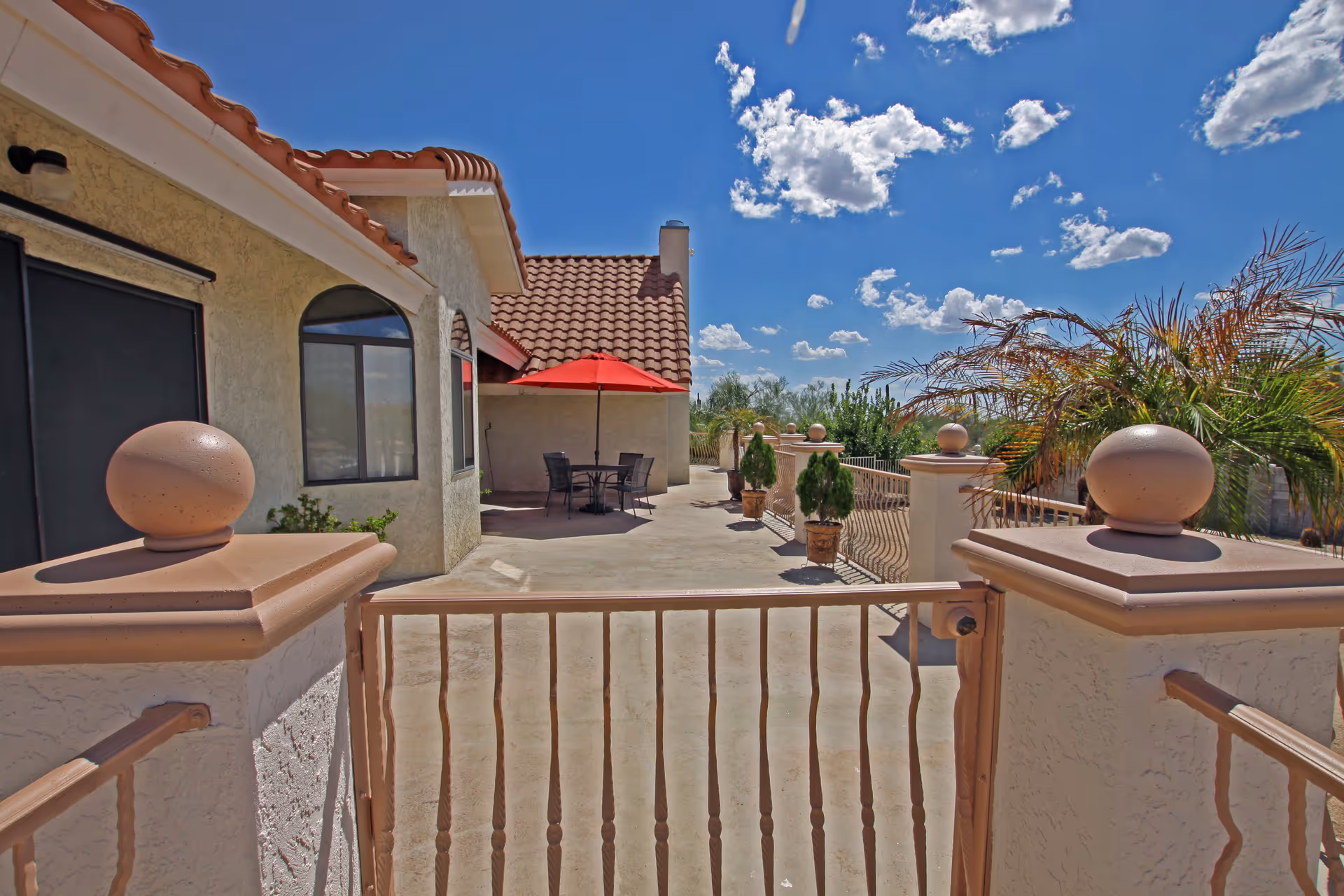 Outdoor patio area with a gated entrance, beige stucco walls, terracotta roof tiles, potted plants along the railing, and a table with chairs under a red umbrella under a blue sky with scattered clouds.