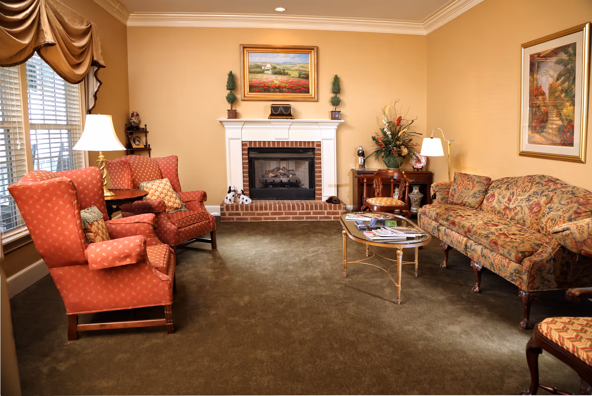 A cozy living room with beige walls and green carpet featuring a brick fireplace in the center. On the left side, there are two red upholstered armchairs and a matching loveseat with patterned cushions near a window with draped curtains. On the right side, there is a floral patterned sofa and a wooden side table with a lamp and a flower arrangement. A glass coffee table with magazines is in the middle of the room. Two framed paintings hang on the walls.