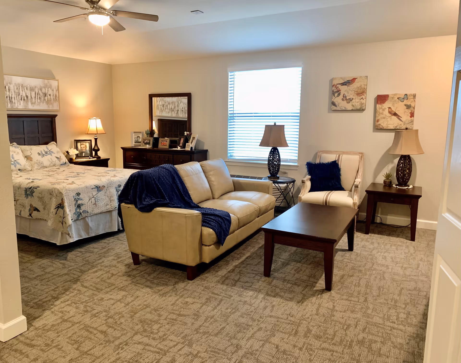 A cozy bedroom with a bed on the left side covered with a floral quilt, a wooden dresser with a mirror and framed photos, a beige leather sofa with a blue throw blanket, a wooden coffee table, a white armchair with a blue cushion, two side tables with lamps, and two bird-themed paintings on the wall. The room has beige walls, carpeted floor, and a ceiling fan with a light.