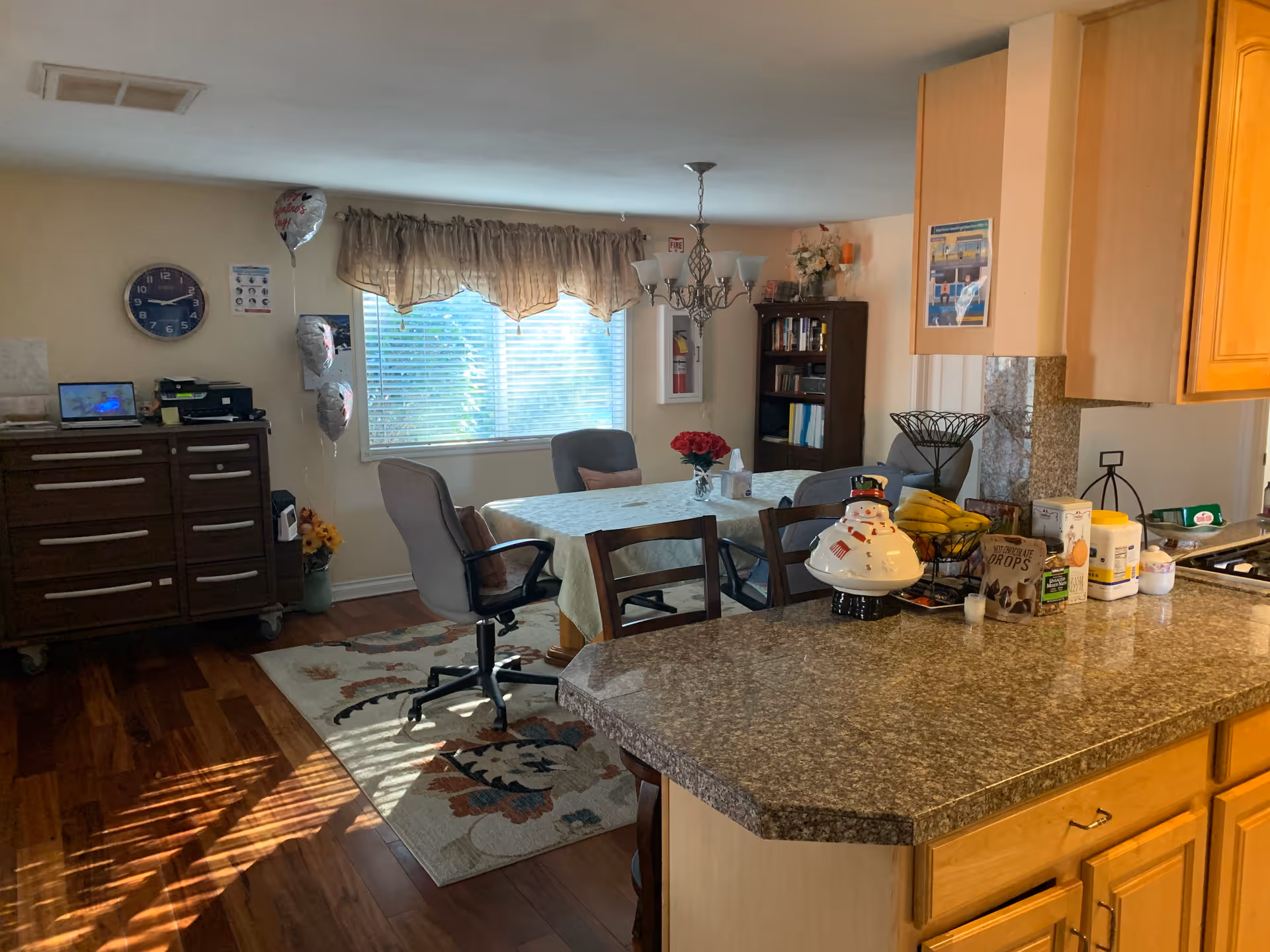 Interior view of a combined kitchen and dining area in a senior living facility. The foreground shows a granite countertop with various items including a snowman cookie jar, bananas, and other kitchen supplies. In the background, there is a dining table covered with a tablecloth, surrounded by chairs, and a window with blinds and a valance letting in natural light. A wooden cabinet with drawers and a clock on the wall are visible to the left, along with some balloons and a small bookshelf in the corner.