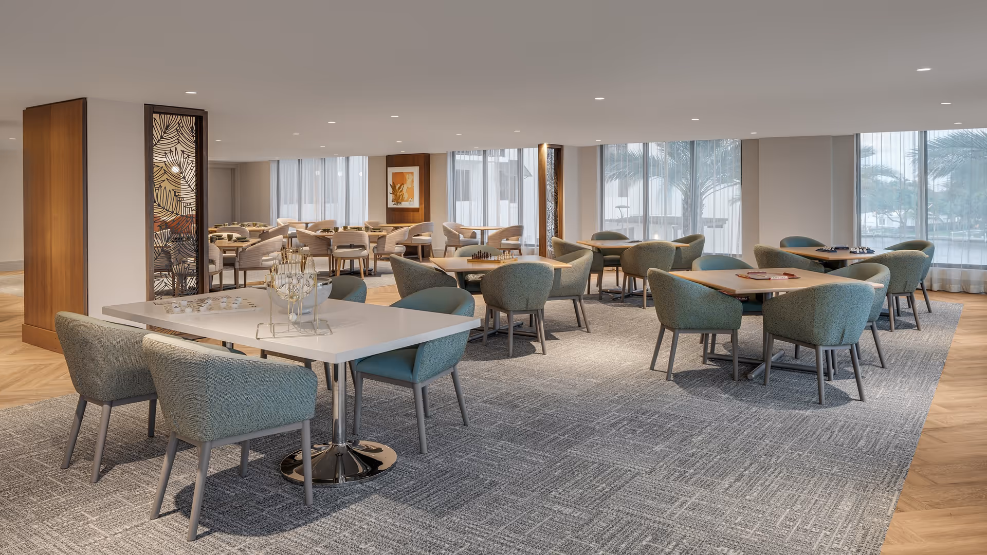 A spacious, well-lit common area with multiple tables and comfortable chairs arranged for socializing or games. The room features large windows with sheer curtains allowing natural light, decorative wooden panels, and a carpeted floor with a light wood border.