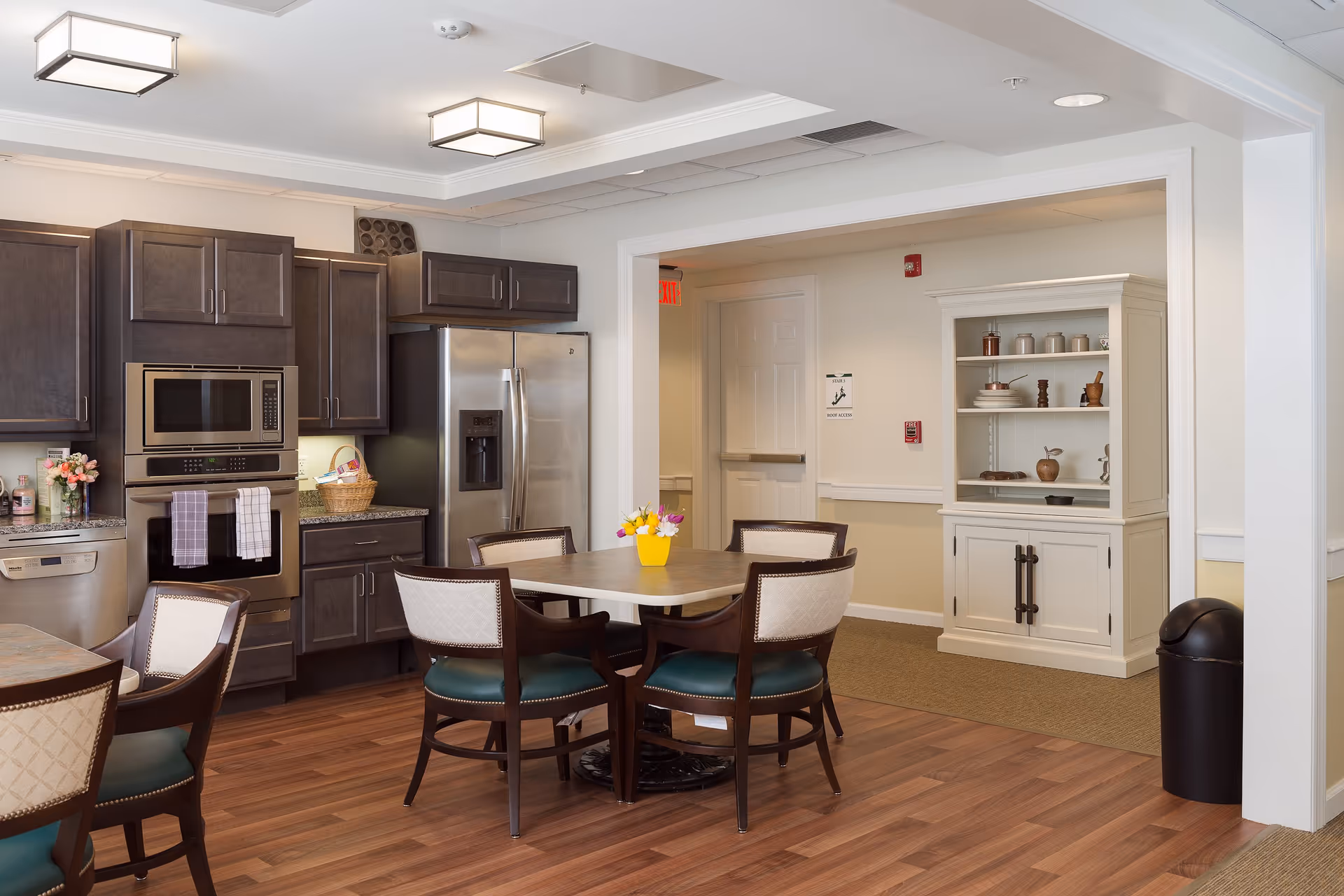 A modern kitchen and dining area in a senior living facility with dark wood cabinets, stainless steel appliances including a refrigerator and microwave, a square dining table with four chairs, and a small yellow vase with flowers on the table. The room has wood flooring, white walls, and ceiling lights. In the background, there is a white cabinet with shelves holding various decorative items and a hallway with an exit sign.