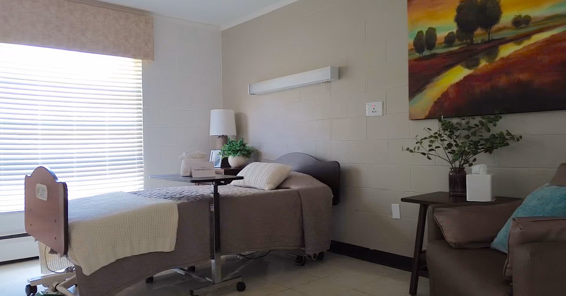 A nursing facility bedroom with a hospital-style bed covered with a beige blanket and pillow. Next to the bed is a small table with a lamp, a plant, and some personal items. A window with closed blinds lets in natural light. On the right side, there is a small table with a tissue box and a vase with greenery, next to a cushioned chair. A colorful landscape painting hangs on the wall above the table.