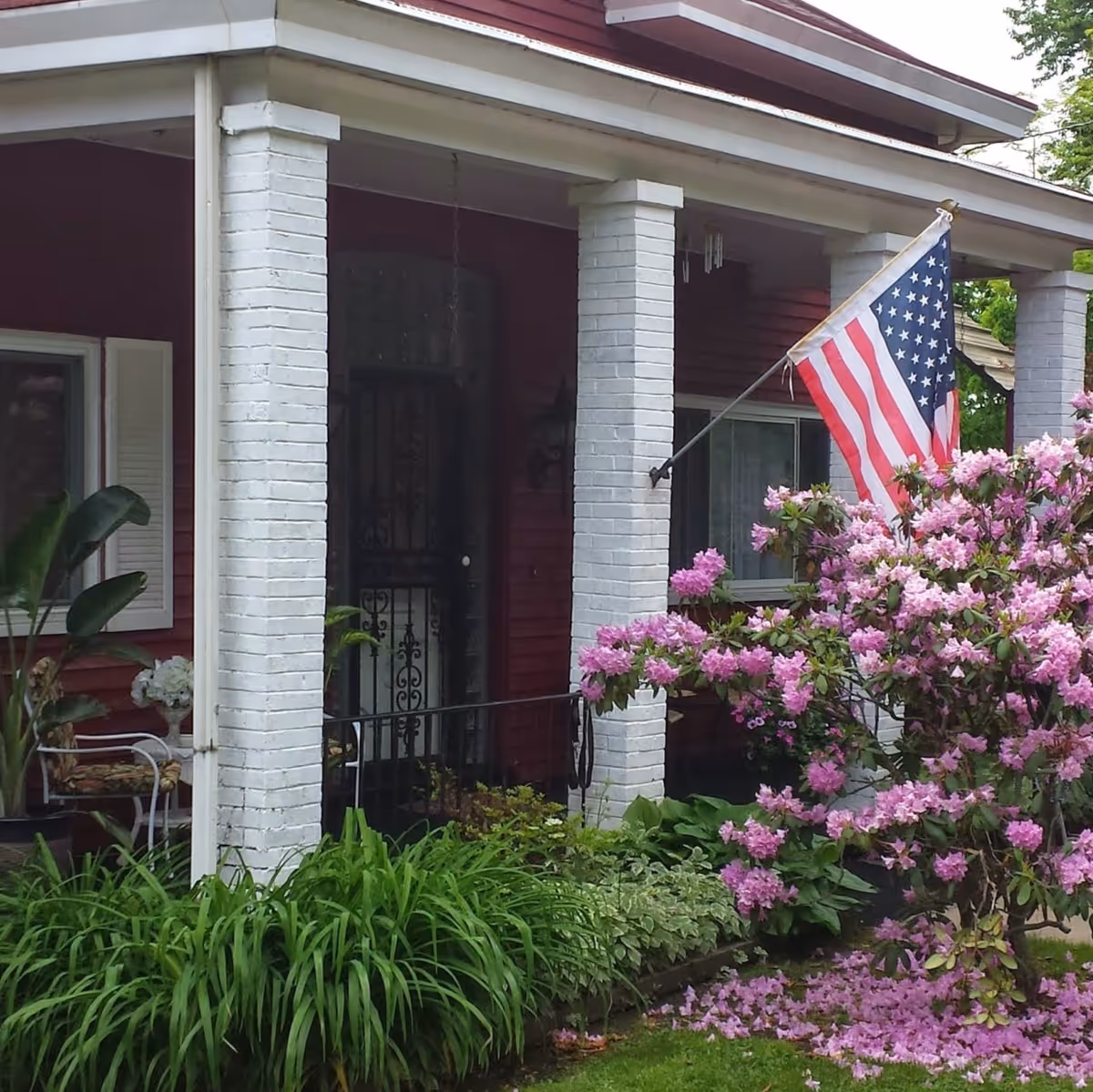 Front porch of a red house with white brick columns, an American flag mounted on one column, and a blooming pink rhododendron bush in the garden in front. There is a black metal security door and some greenery around the porch area.
