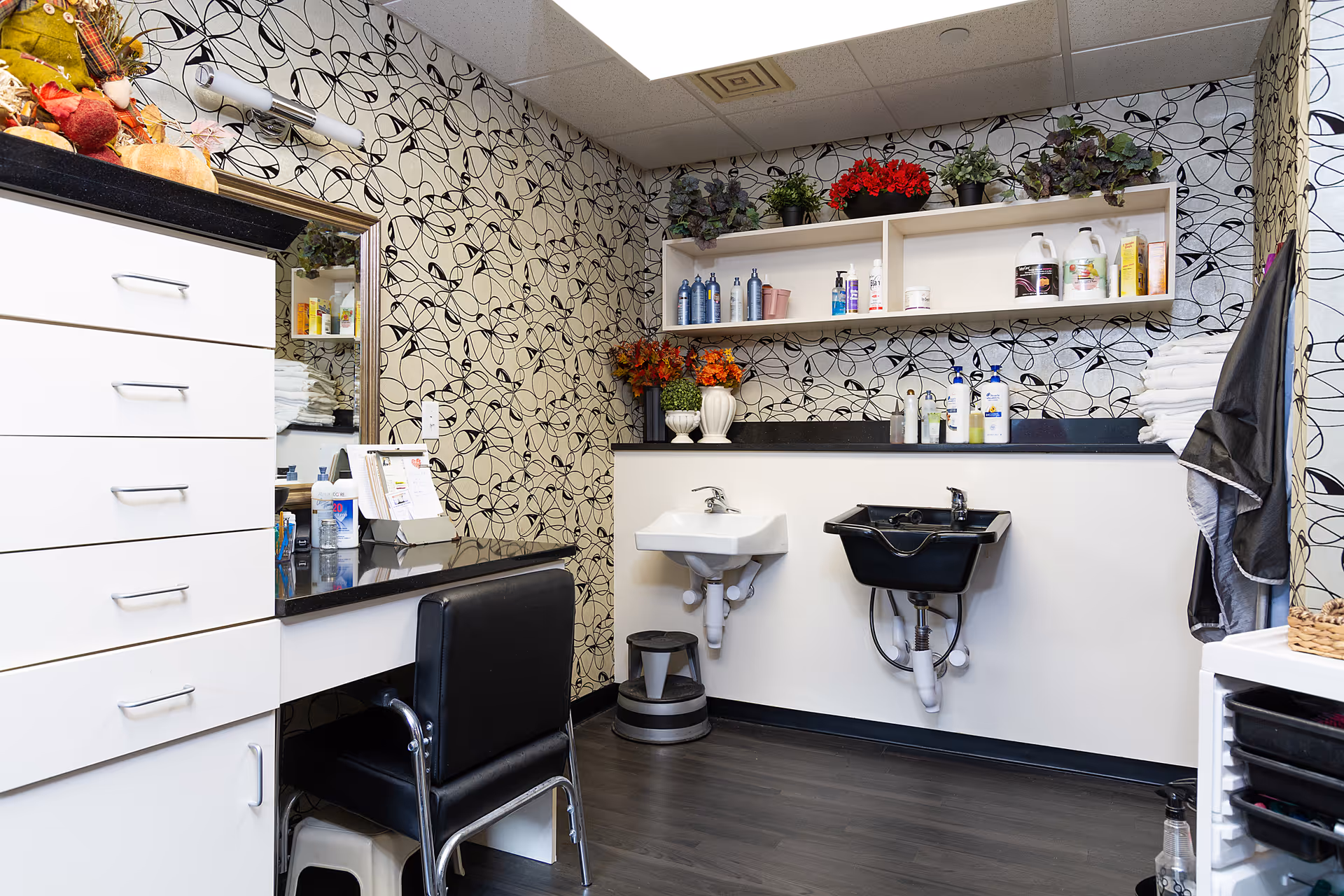 Interior of a salon or grooming area with a black salon chair in front of a counter with drawers. There is a mirror on the wall, patterned wallpaper, a small white sink, and a black shampoo sink. Shelves above hold various bottles and decorative plants. Towels and a hanging cape are visible on the right side.