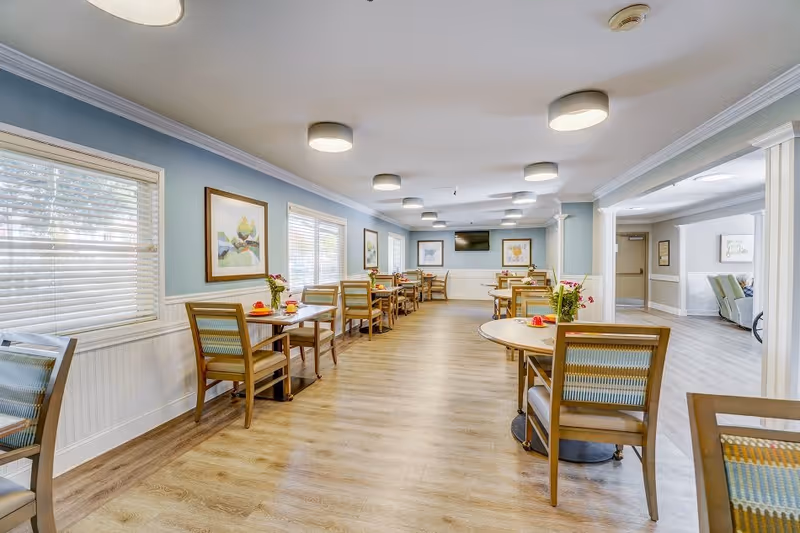 A bright and spacious dining room with wooden tables and chairs arranged neatly. The room has light blue walls with white wainscoting, several windows with blinds, framed artwork on the walls, and ceiling lights. Small flower arrangements and place settings are on the tables.