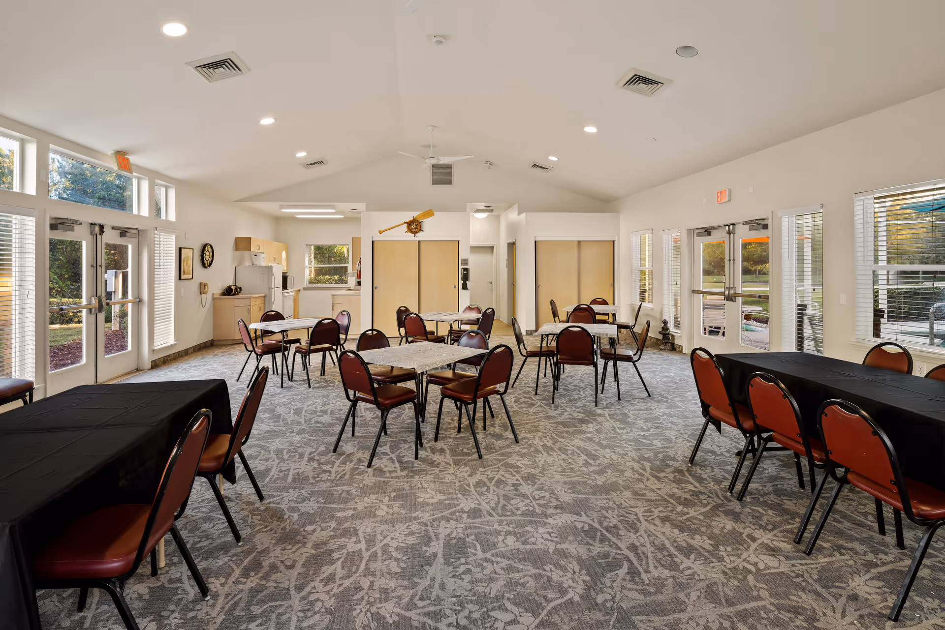 A spacious dining room in a senior living community with several tables and chairs arranged throughout the room. The room has large windows and glass doors allowing natural light to fill the space. There is a small kitchen area with cabinets, a refrigerator, and a microwave in the background. The carpet has a patterned design, and the walls are painted white. Ceiling lights and vents are visible on the ceiling.