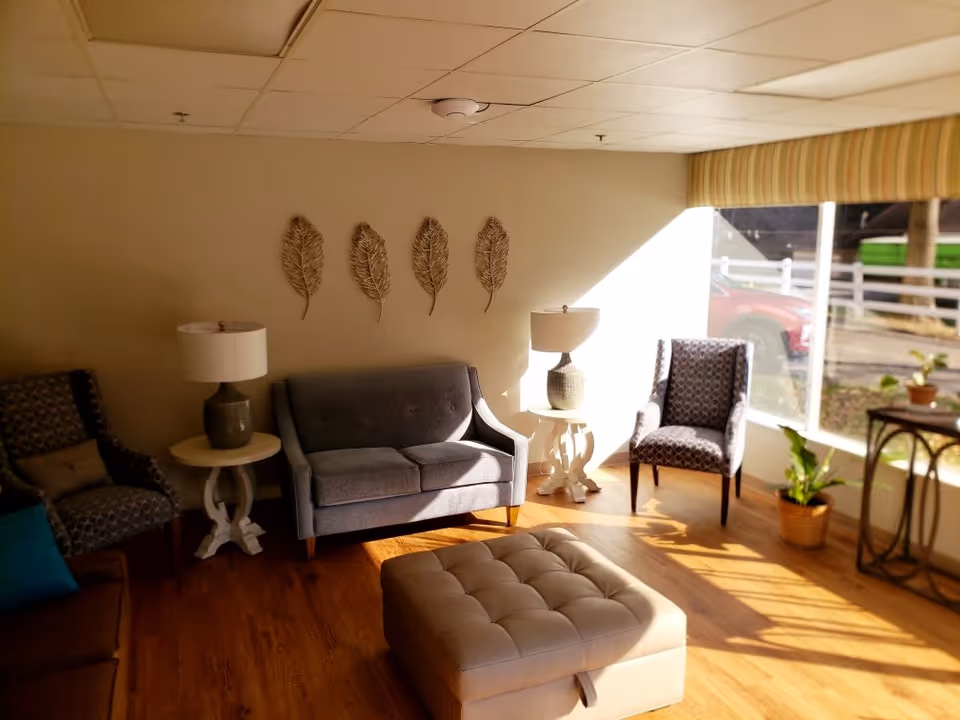 A cozy living room area with wooden flooring, featuring a gray loveseat, two patterned armchairs, a tufted ottoman, two side tables with lamps, and decorative wall art of four metal leaves. Sunlight streams in through a large window with a striped valance, illuminating the room and some indoor plants near the window.