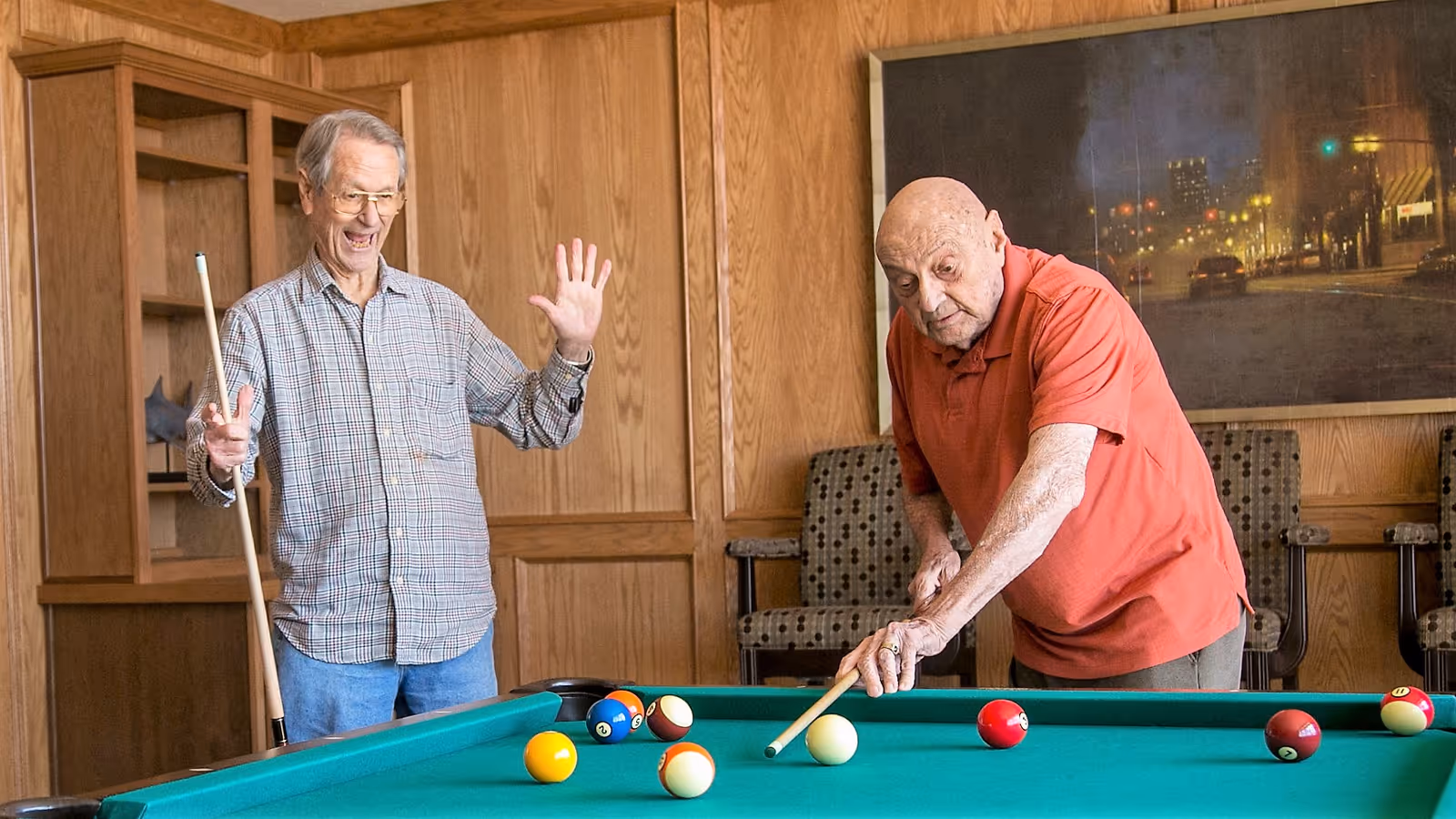 Two elderly men playing pool in a wood-paneled room with chairs and a large framed picture on the wall. One man is aiming with a pool cue while the other watches and gestures with a smile.