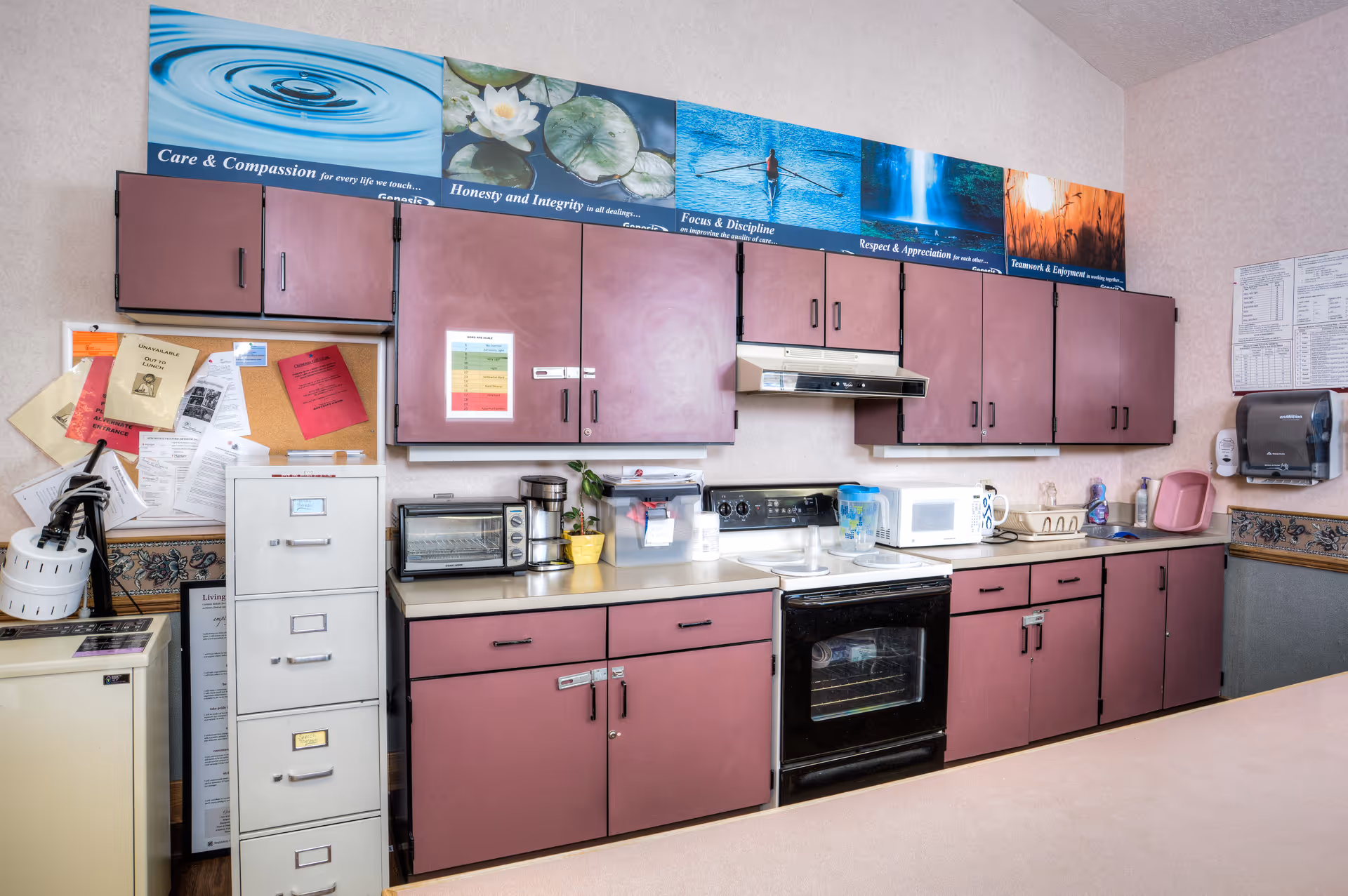 Interior view of a kitchen area in a healthcare facility featuring mauve cabinets, a black stove with an oven, a microwave, a toaster oven, a coffee maker, and various kitchen supplies on the countertops. Above the cabinets are motivational posters with images and text about care, honesty, focus, respect, and teamwork. A filing cabinet and bulletin board with papers are visible on the left side.