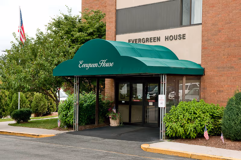 Entrance to Evergreen House Health Center with a green awning displaying the name Evergreen House. The building is made of brick and has glass doors. There are bushes and small American flags near the entrance, and an American flag on a pole to the left.