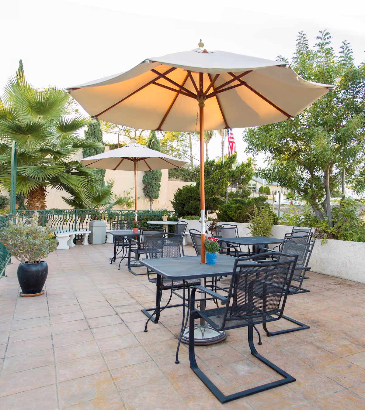 Outdoor patio with metal tables and chairs under large umbrellas surrounded by potted plants and trees.