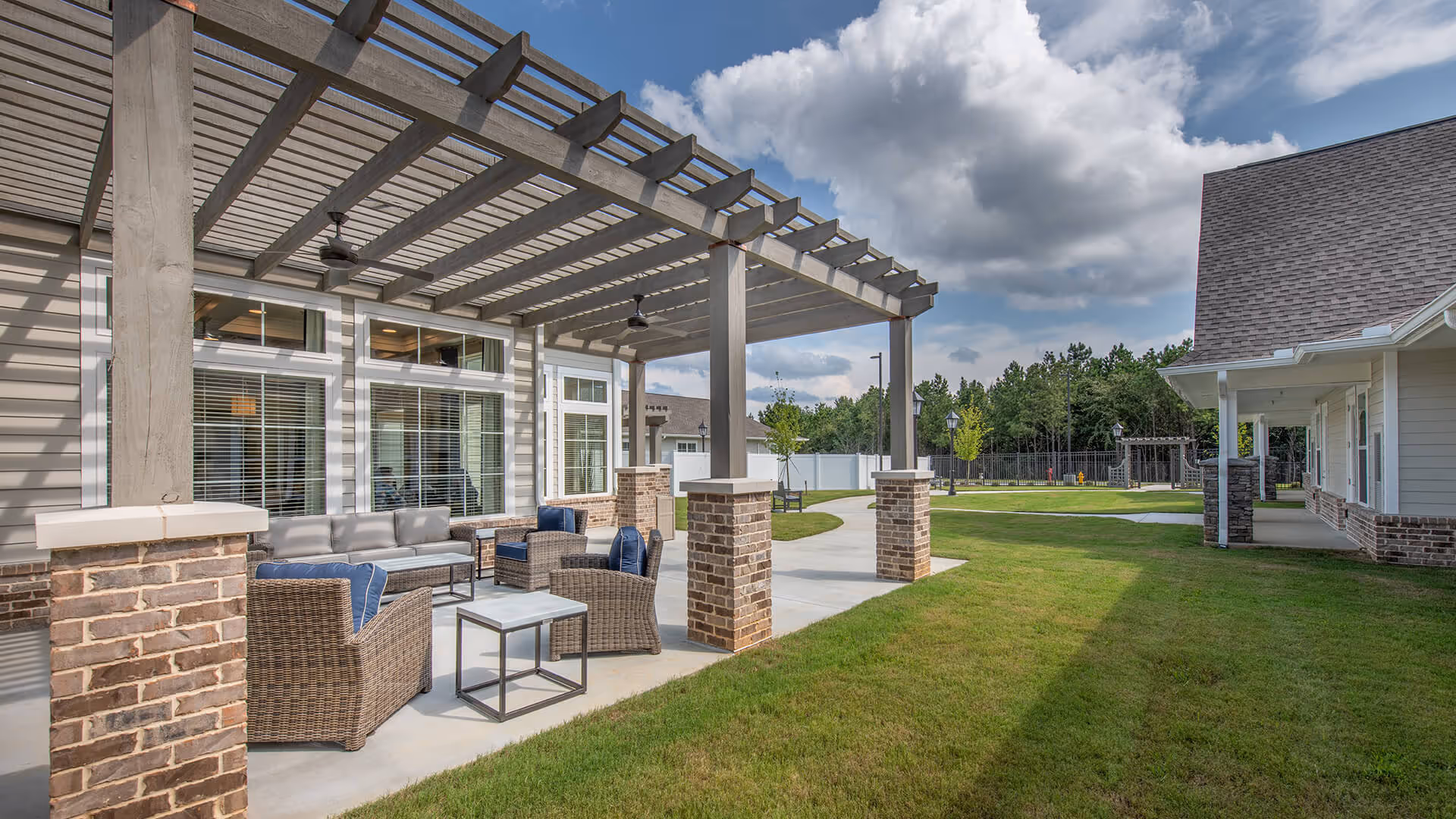 Outdoor covered patio with wicker seating under a pergola beside a senior living building and a grassy courtyard.