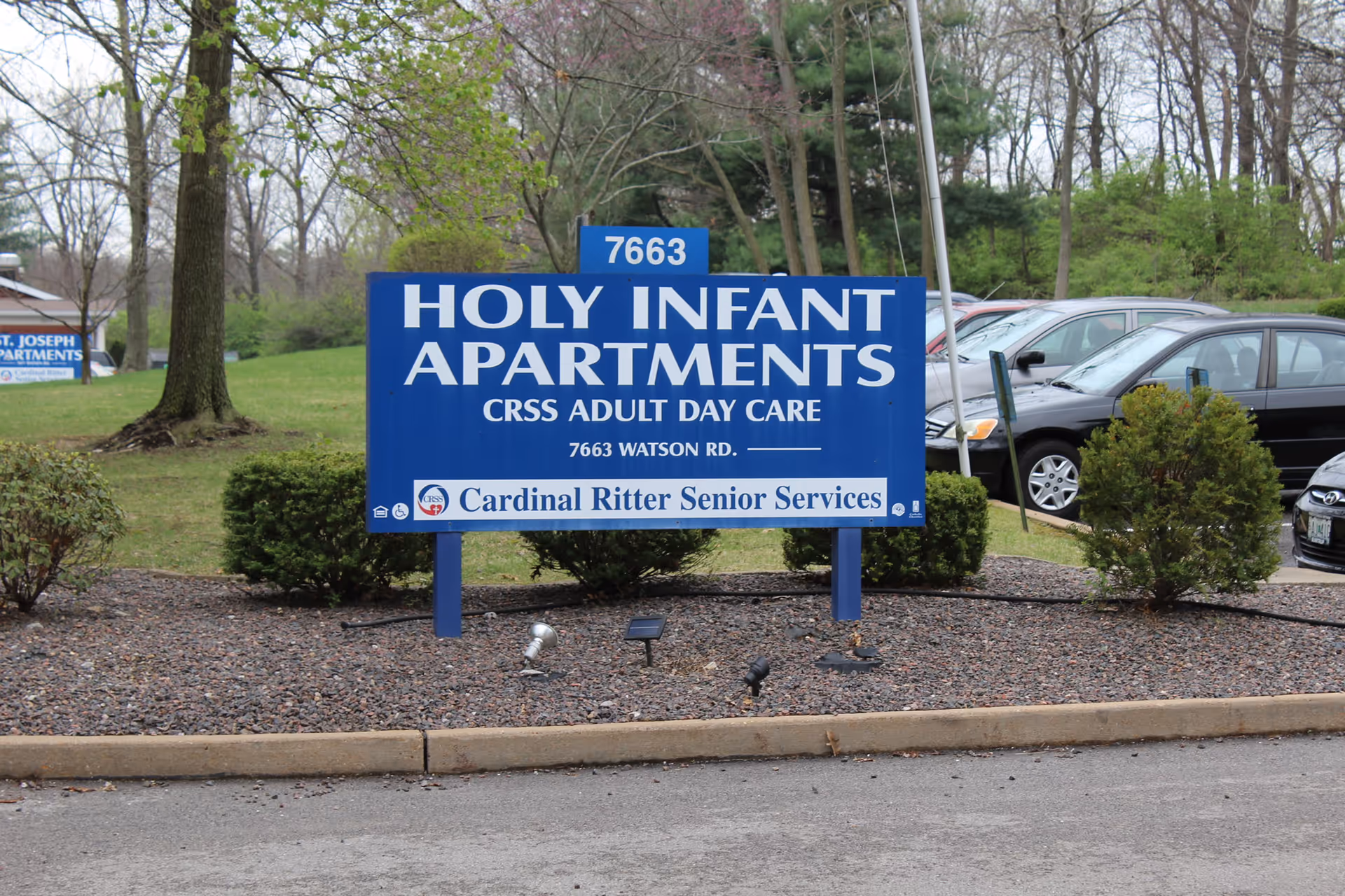 A blue sign for Holy Infant Apartments with information about CRSS Adult Day Care and the address 7663 Watson Rd. The sign also mentions Cardinal Ritter Senior Services. The sign is placed in a landscaped area with bushes and trees, and there are parked cars visible in the background.