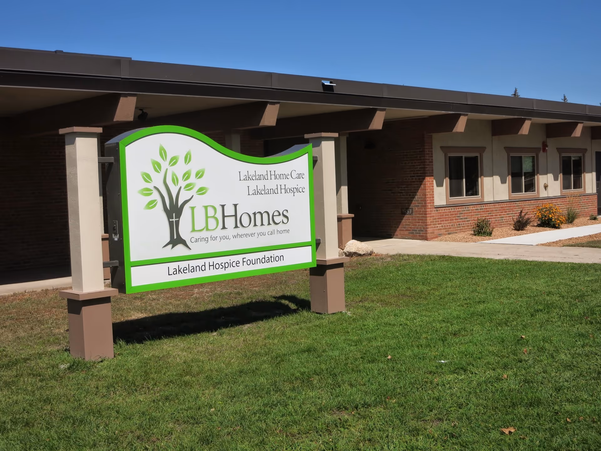 Outdoor view of a single-story building with a sign in front that reads 'Lakeland Home Care Lakeland Hospice LB Homes Caring for you, wherever you call home Lakeland Hospice Foundation'. The building has brick and beige walls with several windows and a small garden area with flowers.
