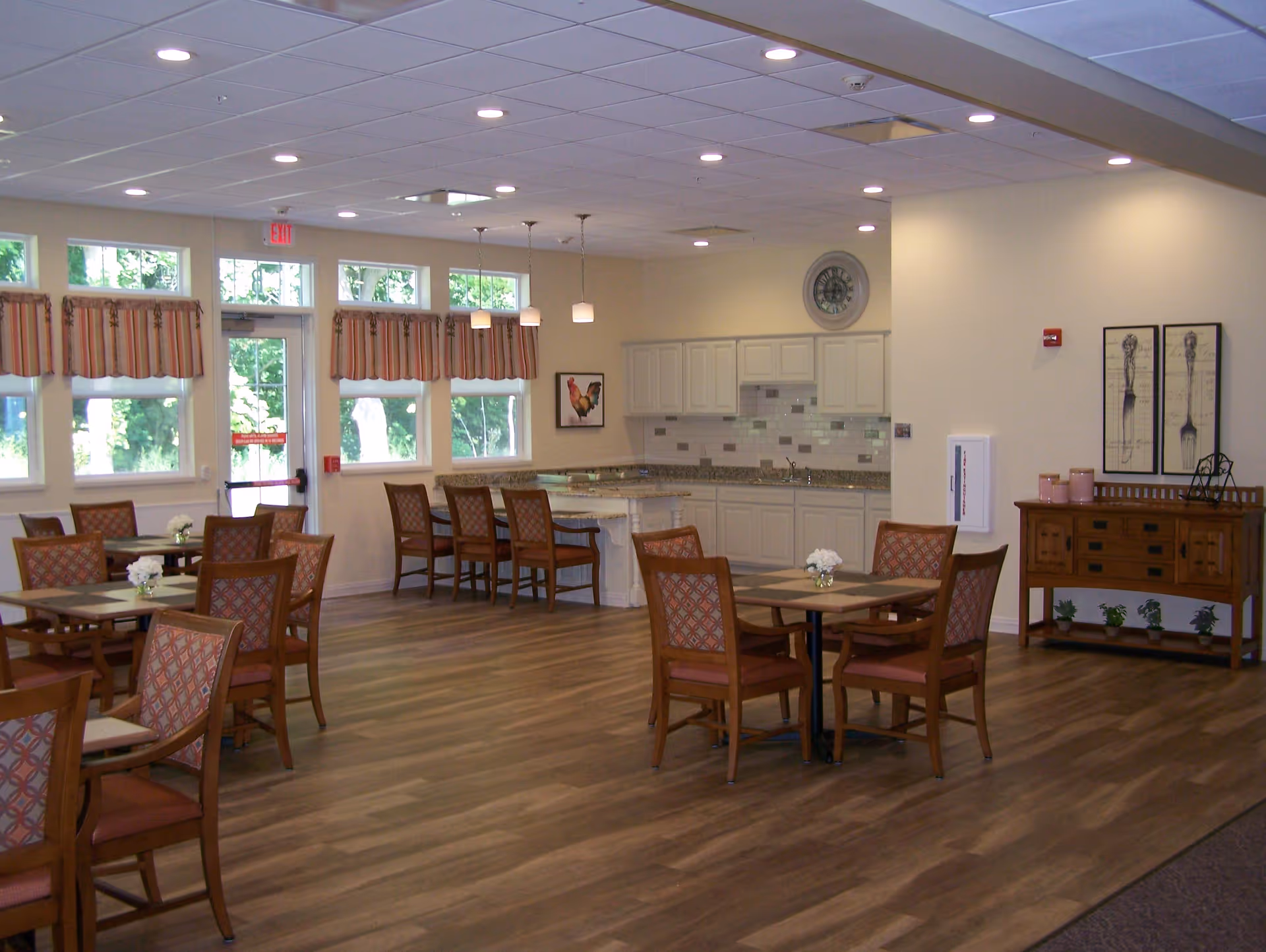 A spacious dining area with several wooden tables and chairs arranged neatly. The room features large windows with striped valances allowing natural light to enter. In the background, there is a kitchen area with white cabinets, a granite countertop, and a tiled backsplash. The floor is wood-style laminate, and the ceiling has recessed lighting and hanging pendant lights. Decorative wall art and a wooden sideboard with plants and candles are also visible.