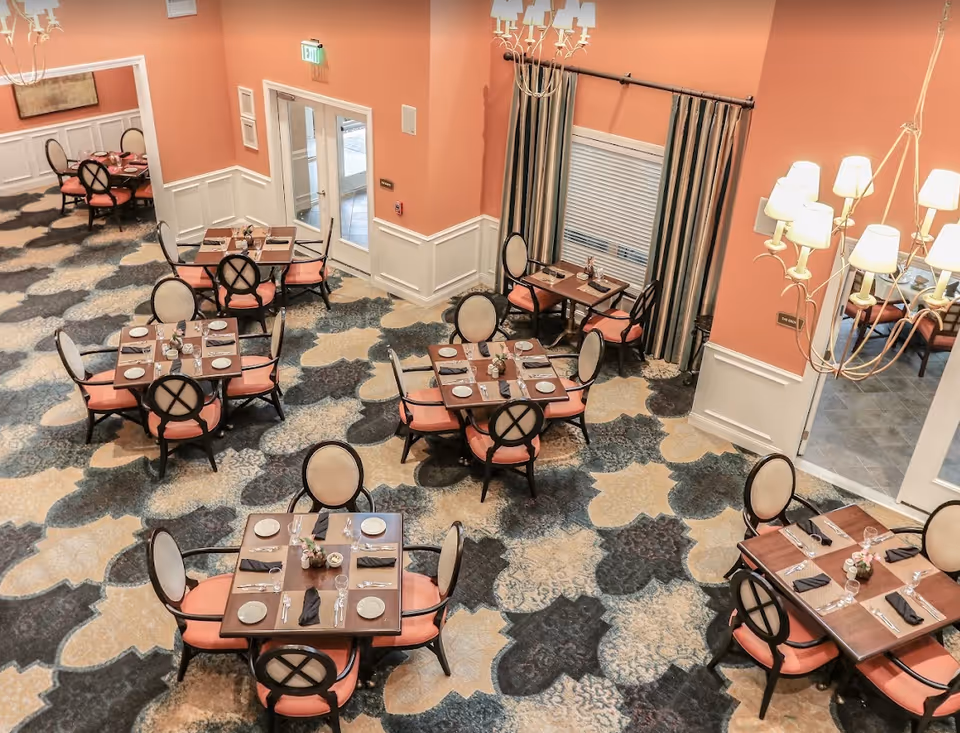 Interior view of a dining room with multiple square wooden tables set with plates, glasses, and cutlery. Each table is surrounded by four chairs with peach-colored cushions and oval backs. The room has peach-colored walls with white wainscoting, patterned carpet in shades of blue, beige, and gray, and large windows with striped curtains. A chandelier with multiple lampshades hangs from the ceiling.