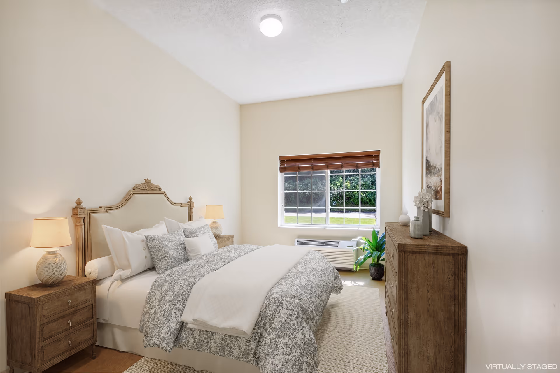 A neatly arranged bedroom with a large bed featuring multiple pillows and a patterned comforter. There are two wooden nightstands with lamps on either side of the bed. A wooden dresser with decorative items and a framed picture is positioned against the right wall. A window with wooden blinds allows natural light to brighten the room, and a small green plant sits near the window.