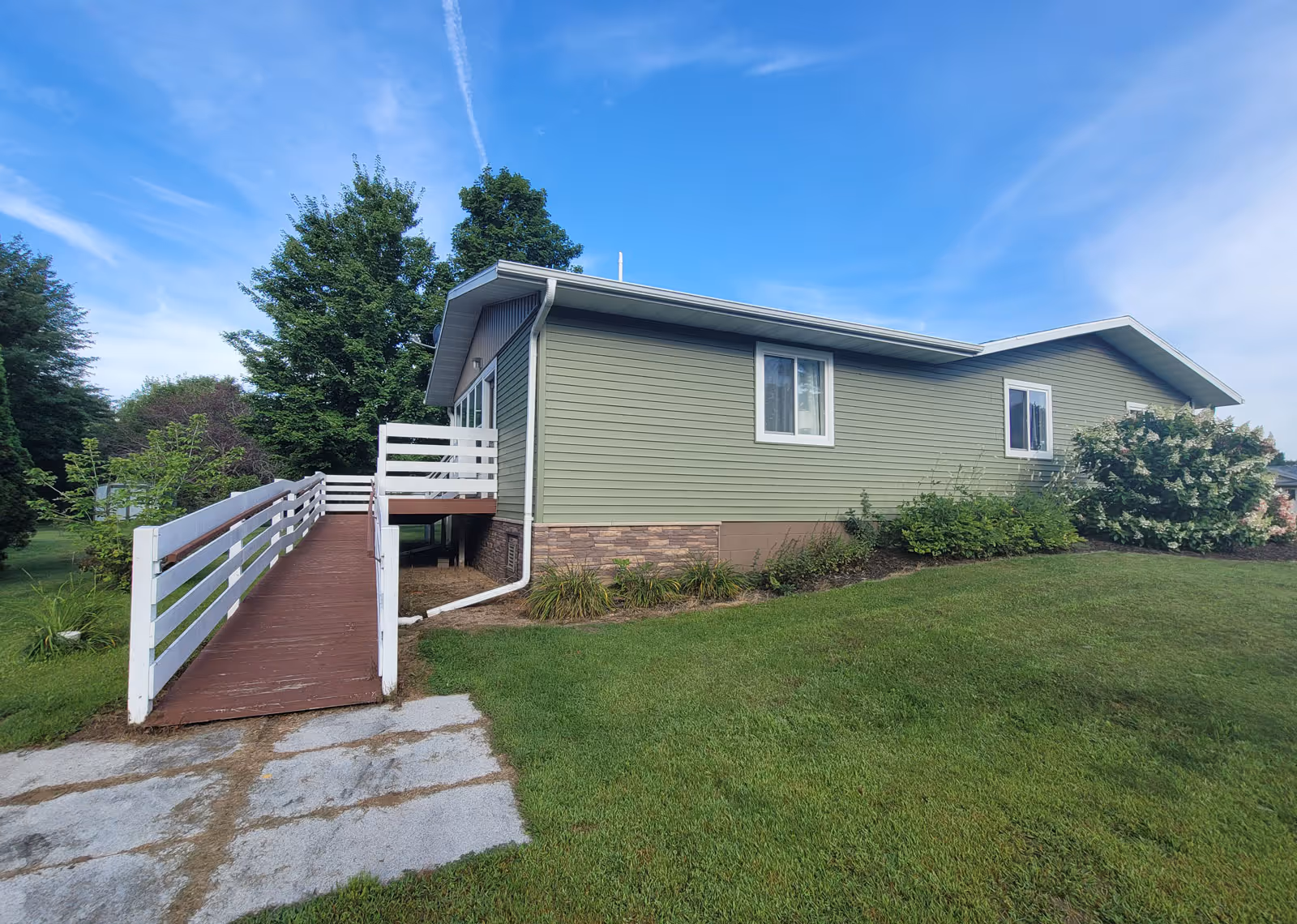 Exterior view of a single-story green building with white trim, featuring a wooden ramp with white railings leading to the entrance. The building is surrounded by green grass, bushes, and trees under a blue sky with some clouds.