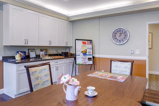 Communal dining area with a wooden table and chairs, a kitchenette with white cabinets, a wall clock, and a bulletin board.