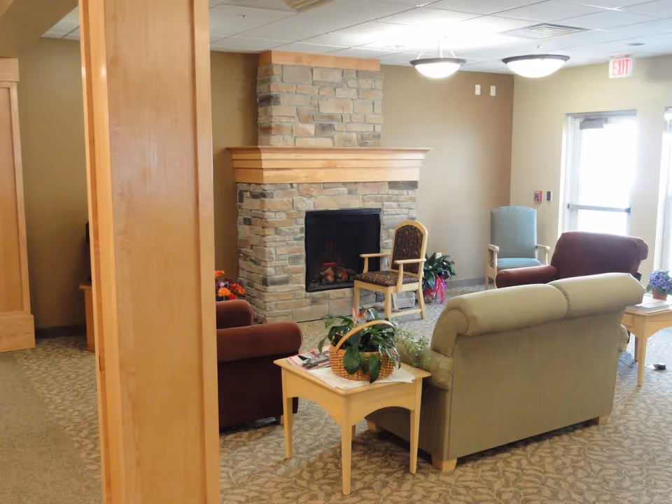 A cozy living room area in a senior living facility featuring a stone fireplace with a wooden mantle, several upholstered chairs and sofas arranged around small wooden tables, potted plants, and soft lighting from ceiling fixtures. There is an exit door with a glass window letting in natural light.