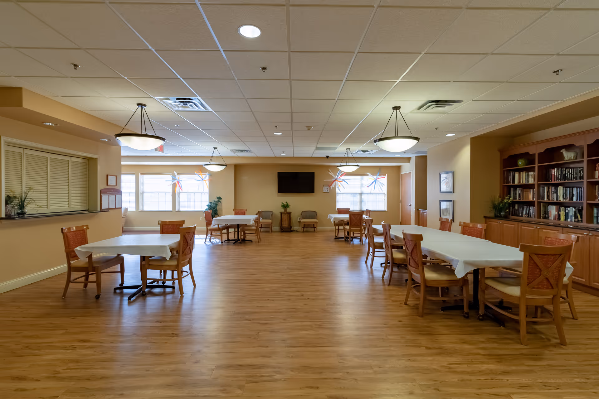 A spacious dining room with several tables covered with white tablecloths and wooden chairs arranged around them. The room has wooden flooring, beige walls, and a ceiling with recessed lights and hanging light fixtures. There are large windows with decorative star-shaped ornaments, a flat-screen TV mounted on the far wall, and a wooden bookshelf filled with books on the right side.