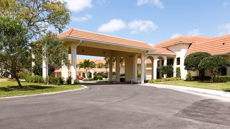 Front entrance of a single-story senior living building with a covered porte-cochere, tiled roof, driveway, and landscaped grounds.