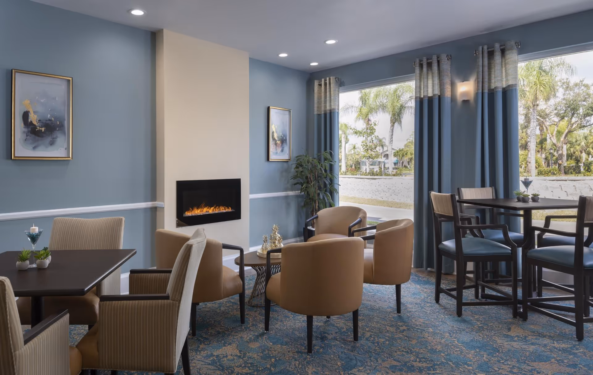 Bright communal lounge and dining area with chairs and tables around a modern fireplace and large windows showing palm trees outside.