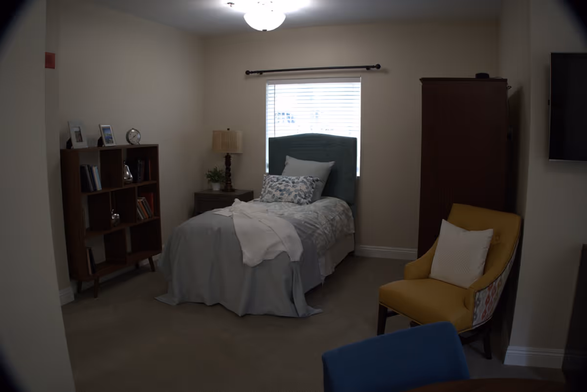 A furnished bedroom with a single bed, bedside table and lamp, bookshelf, wardrobe, and an upholstered chair.