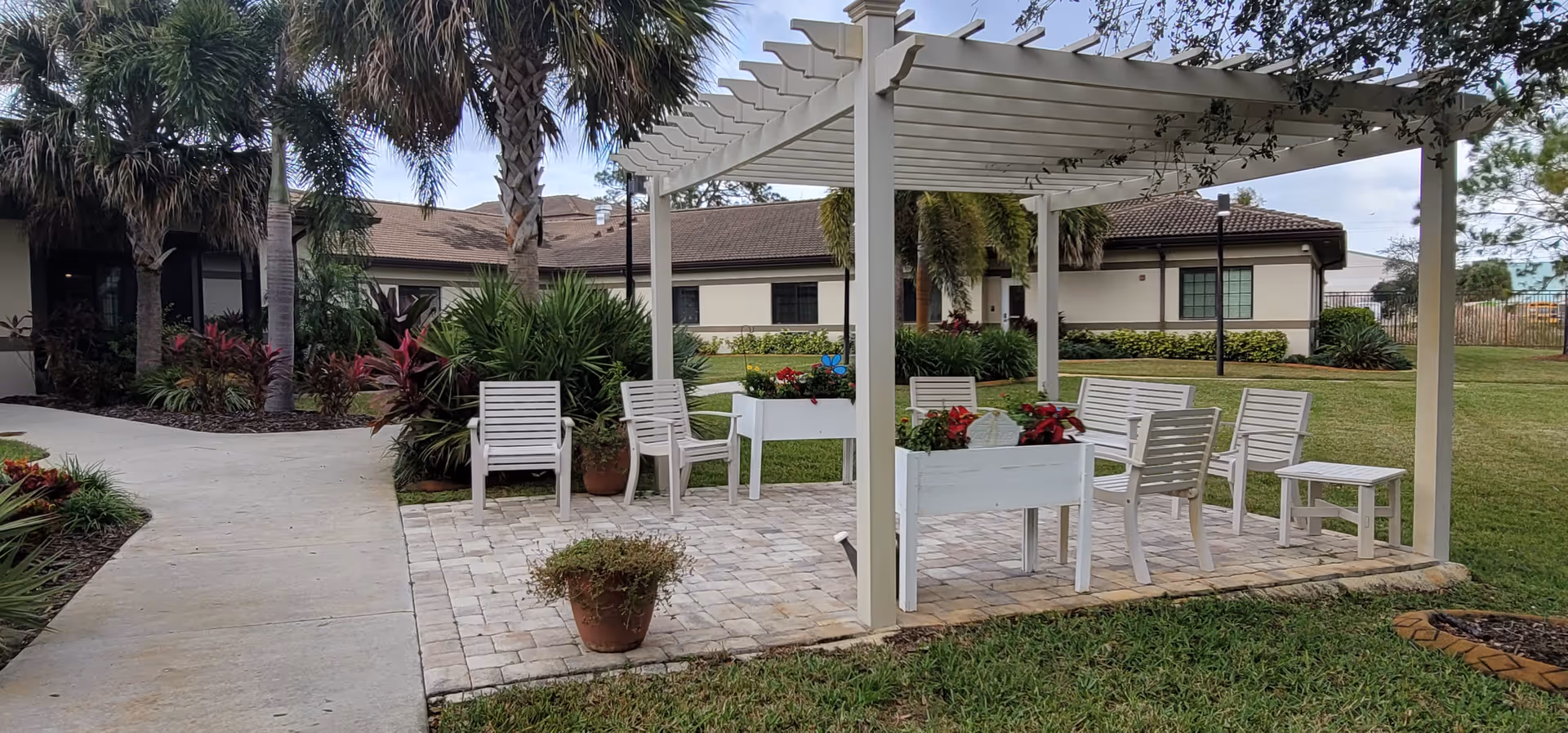 Outdoor patio area with a white pergola covering several white chairs and planter boxes with red flowers. The patio is paved with stone tiles and surrounded by grass and tropical plants, including palm trees. A beige building with a brown roof is visible in the background under a partly cloudy sky.