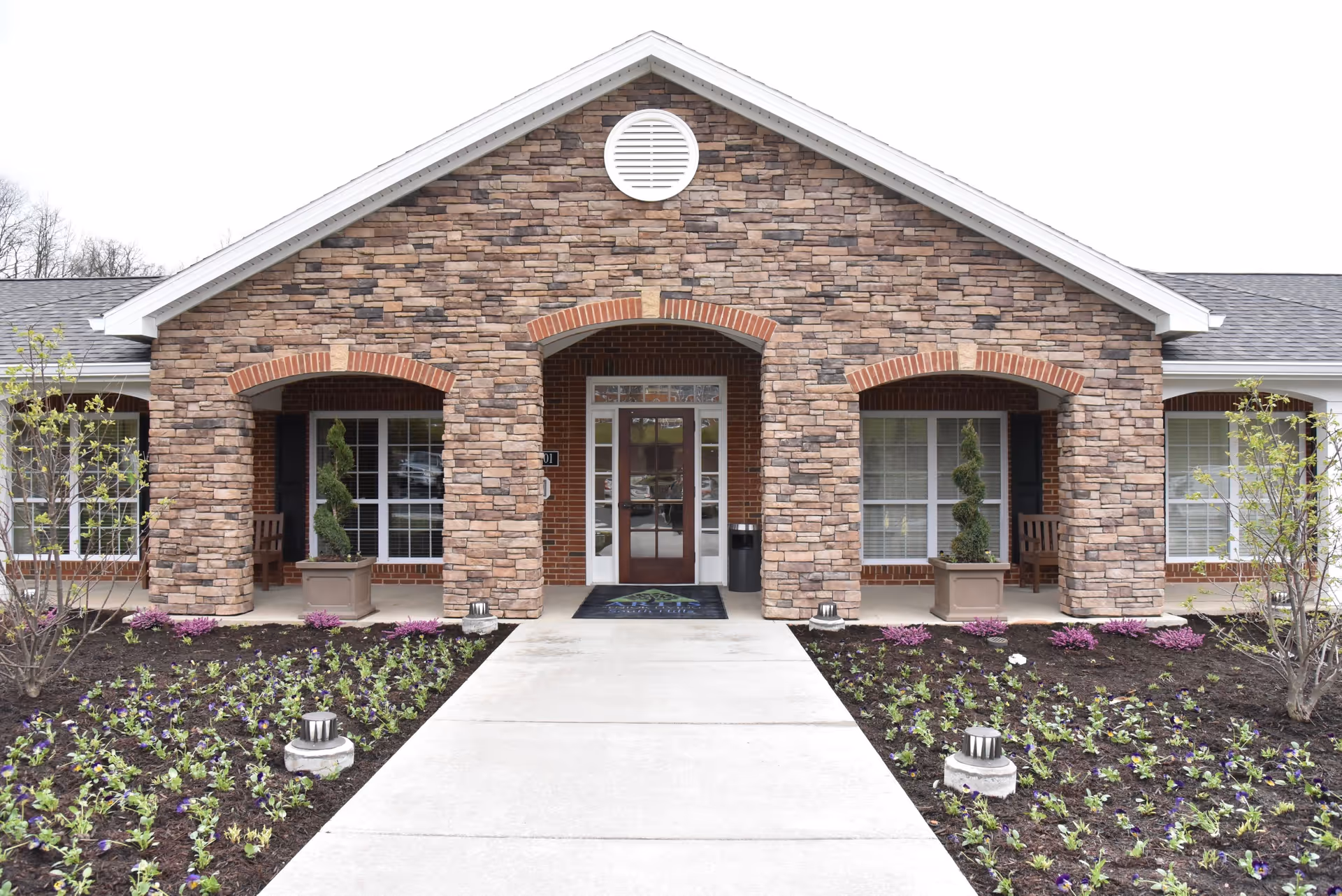 Front exterior view of a senior living facility with a stone and brick facade, a central entrance door with glass panels, two potted topiary plants on either side of the entrance, and flower beds lining the walkway leading to the door.