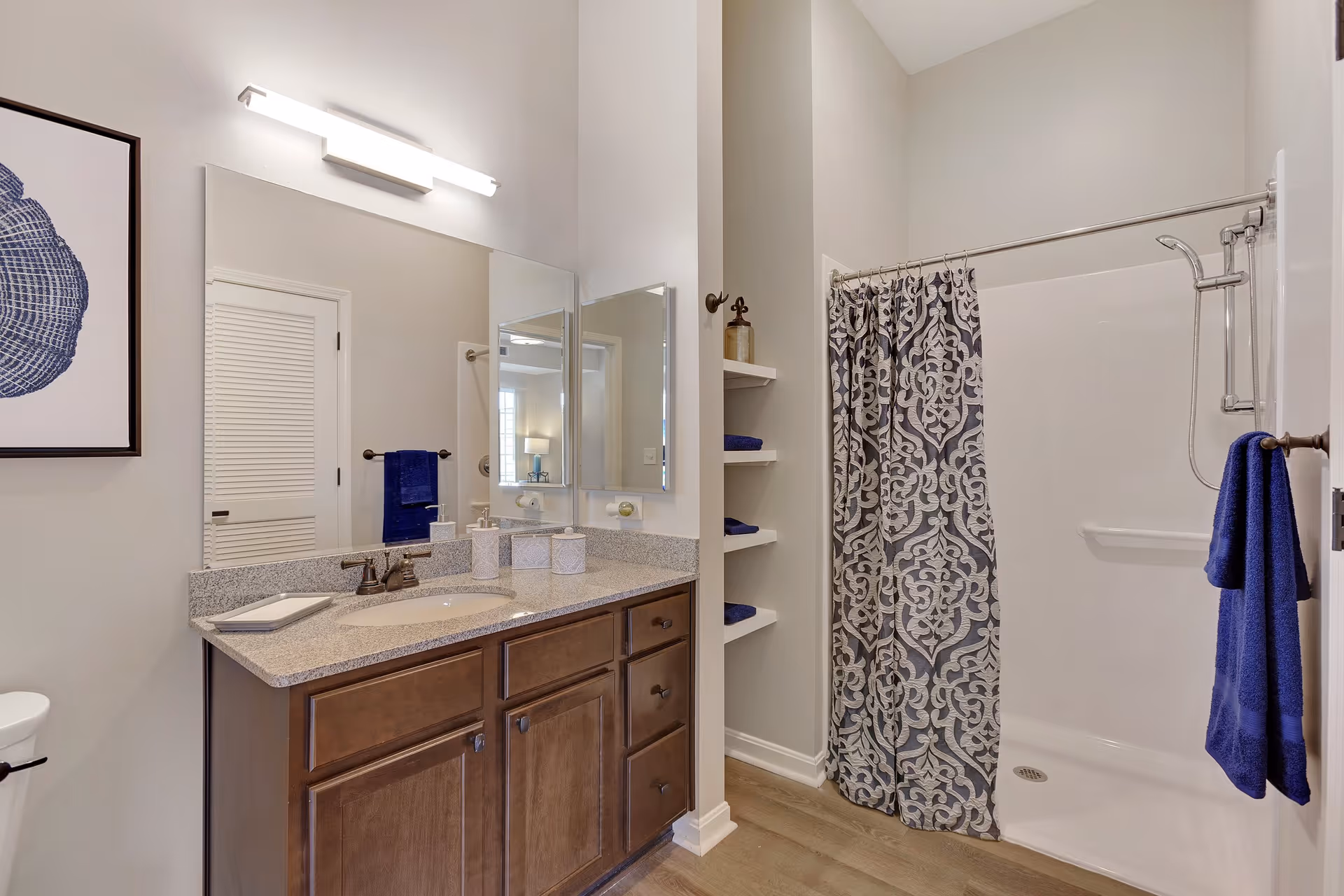 A bathroom with a wooden vanity featuring a granite countertop, a sink with a faucet, and three decorative containers. Above the vanity is a large mirror with a modern light fixture. To the right is a shower area with a patterned shower curtain and a blue towel hanging on a hook. There are built-in shelves with folded blue towels and a framed artwork on the left wall.