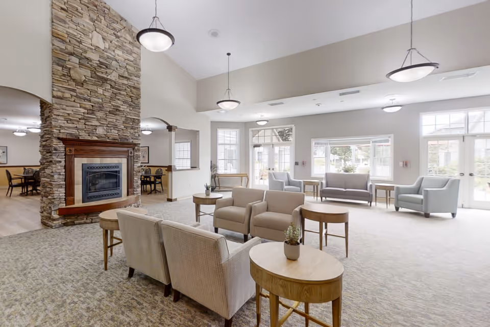 A spacious and well-lit living room area in a senior living facility with multiple armchairs and sofas arranged around small wooden tables. There is a large stone fireplace on the left side, and large windows and glass doors letting in natural light. The room has a neutral color palette with beige and light gray tones.