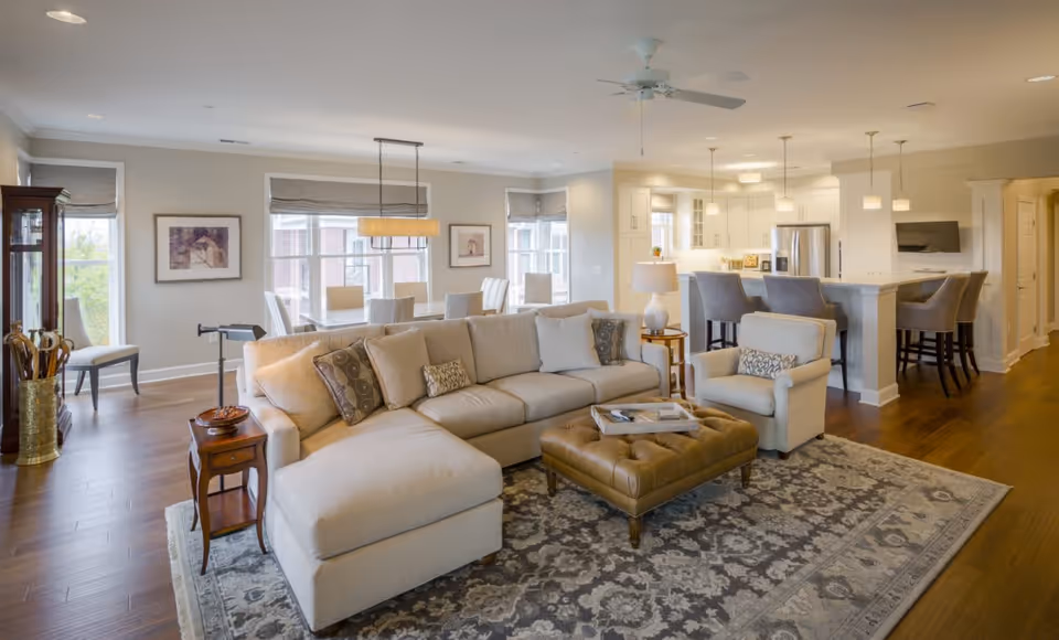 A spacious and well-lit living area featuring a large beige sectional sofa with patterned cushions, a matching armchair, and a tufted leather ottoman on a decorative rug. Behind the sofa is a dining area with a table and chairs near large windows with roman shades. The open kitchen includes a breakfast bar with stools, white cabinetry, stainless steel appliances, and pendant lighting. Hardwood floors and neutral wall colors create a warm and inviting atmosphere.