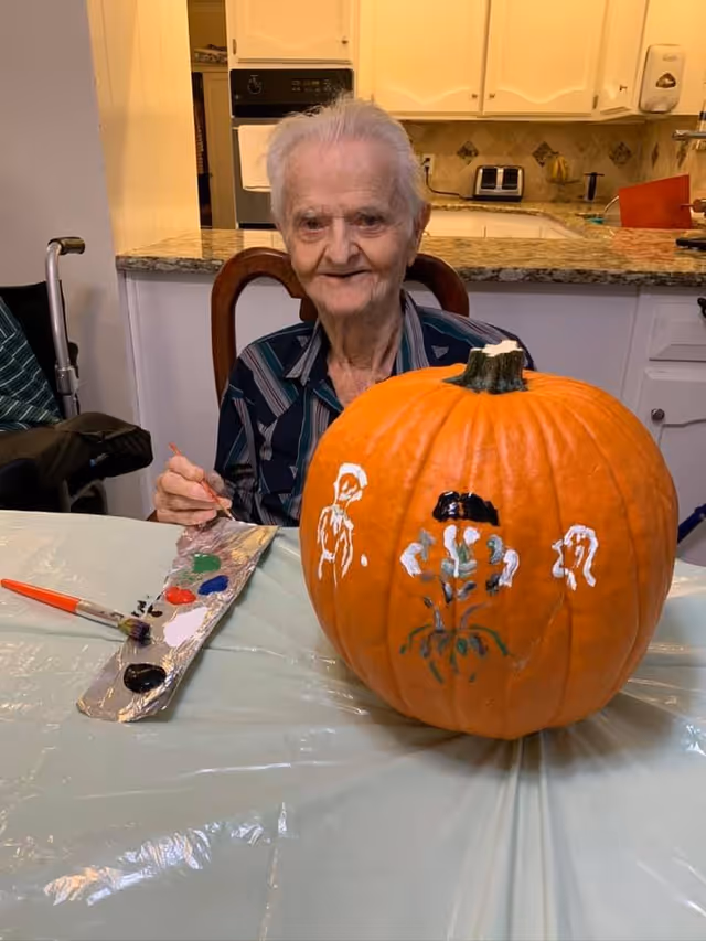 An elderly person sitting at a table in a kitchen area, painting a large pumpkin with various colors. The pumpkin has white, black, and green painted designs on it. The person is smiling and holding a paintbrush, with a paint palette on the table.
