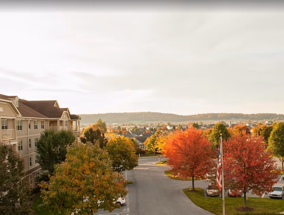 View of a senior living facility parking lot with colorful autumn trees, an American flag on a flagpole, and a multi-story beige building on the left side. The background shows a residential neighborhood and rolling hills under a partly cloudy sky.