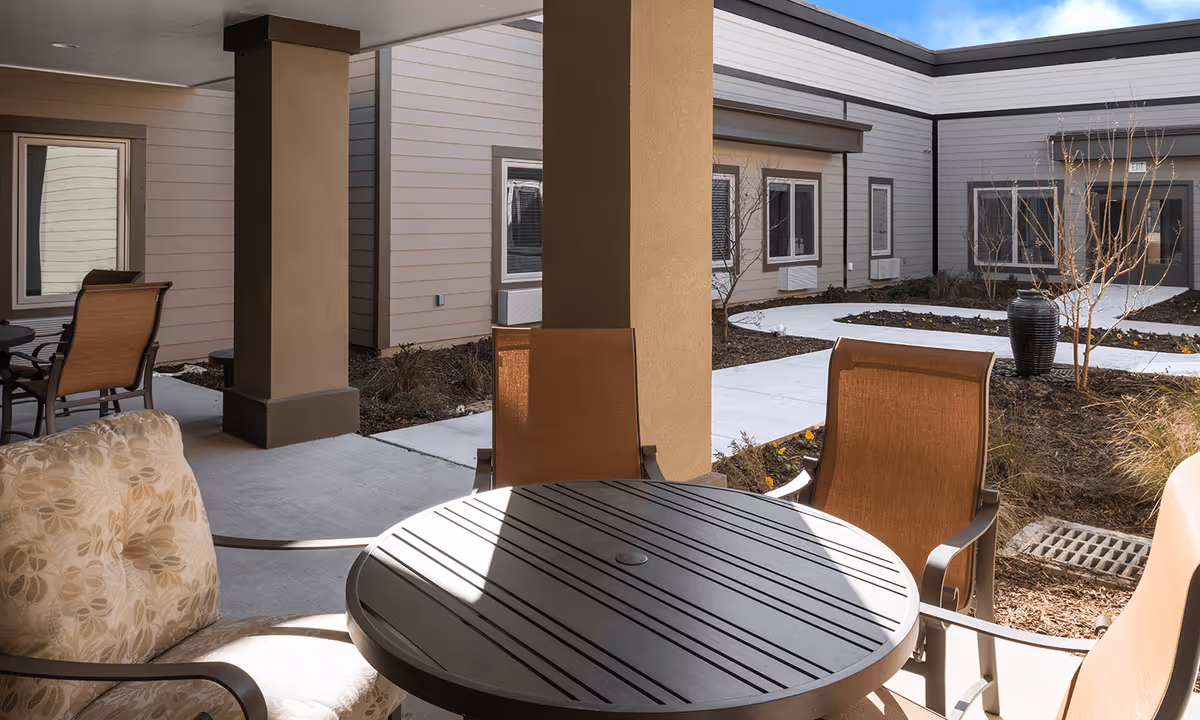 Covered patio with a round table and chairs overlooking a landscaped courtyard and surrounding building exterior.