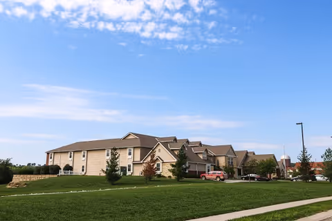 Exterior view of a large, beige senior living facility building with multiple roof peaks, surrounded by green grass and a few small trees under a blue sky with scattered clouds. There are a few cars parked near the entrance.