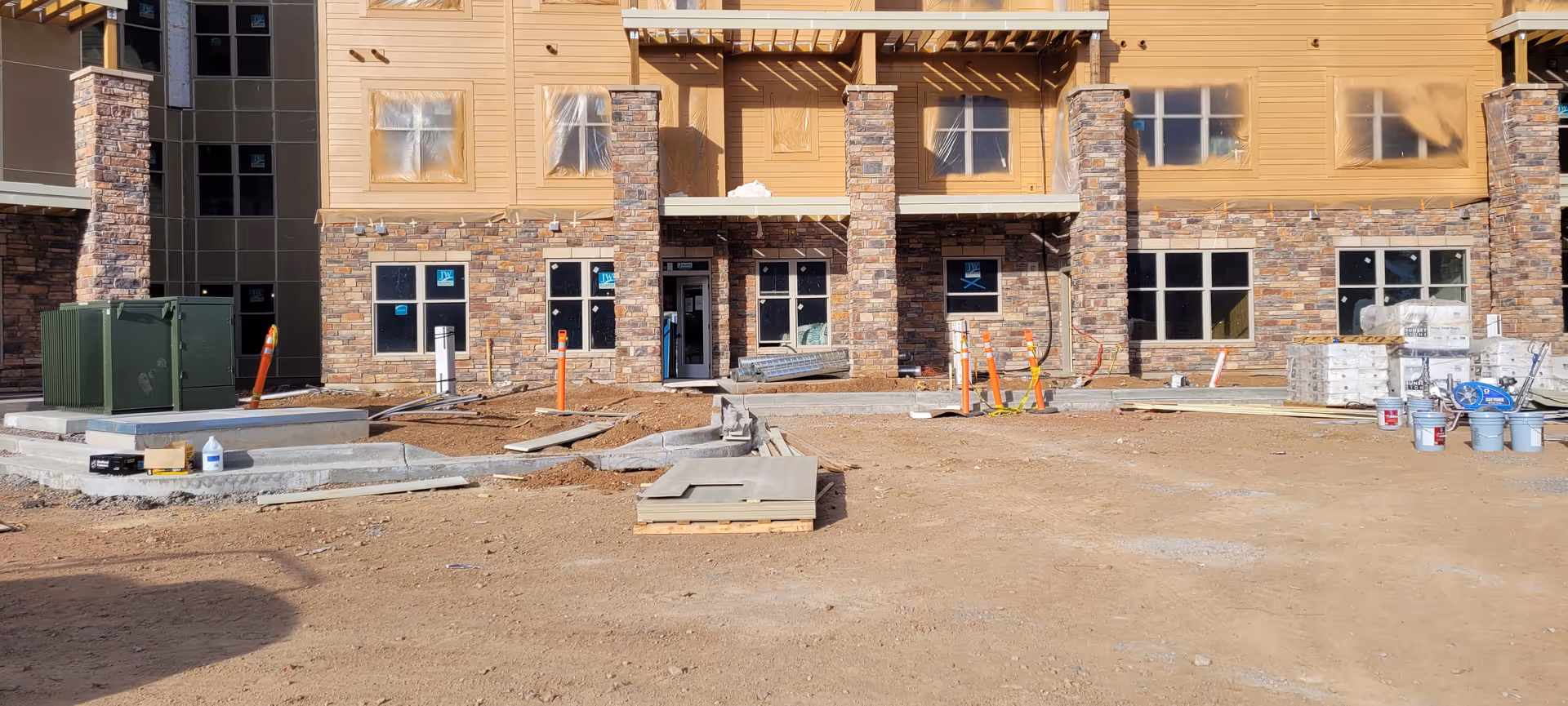 Front exterior view of a building under construction with stone and wood siding, multiple windows, and several construction materials and equipment scattered around the dirt ground in front.