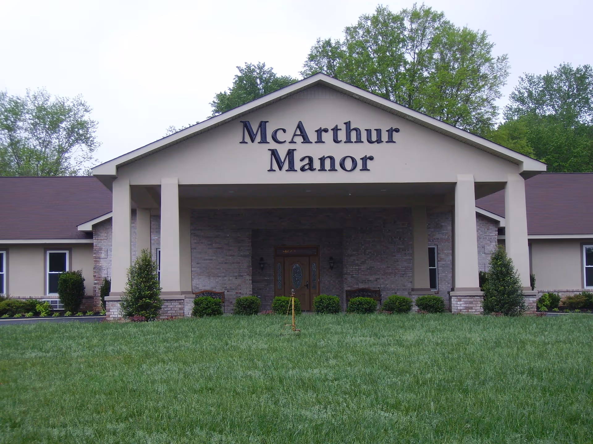 Front exterior view of McArthur Manor Assisted Living building with a covered entrance, beige walls, brick accents, and a well-maintained green lawn in front.